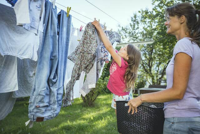 Jente med langt, blondt hår, knall rosa t-skjorte og mønstrete hvit shorts og fester klær med klesklyper på ei tørkesnor. Bak henne står ei kvinne med langt mørkt hår, lyse lilla t-skjorte og jeans, og holder en kurv med klær. Foto Gerhard Berger