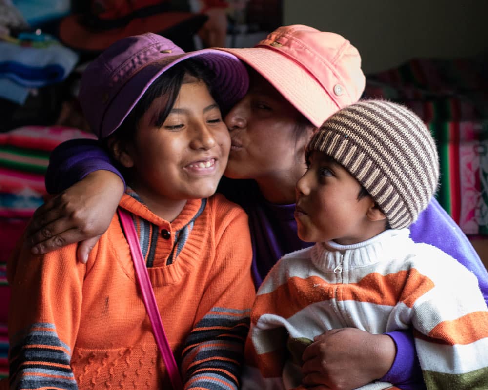 Mor fra Peru med lys oransje hatt har begge barna på fanget. Hun kysser datteren som har på seg lilla hatt og oransje genser, sønnen titter opp på dem han har stripete strikkelue og oransje og hvit stripet genser. Familien har fått støtte gjennom vårt forebyggende arbeid. Foto: Alejandra Kaiser