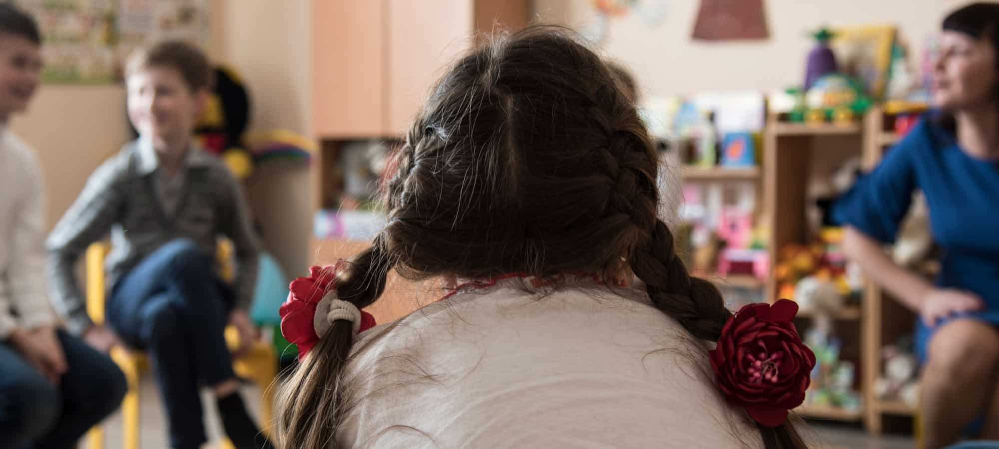 Flere barn sitter i et rom med leker og snakker med psykologen. Ei jente, med fletter og hvit genser, sitter på en stol i forgrunnen med ryggen til. Psykologer er en del av SOS-barnebyers team i områder med krig og konflikt. Foto: Katerina Ilievska