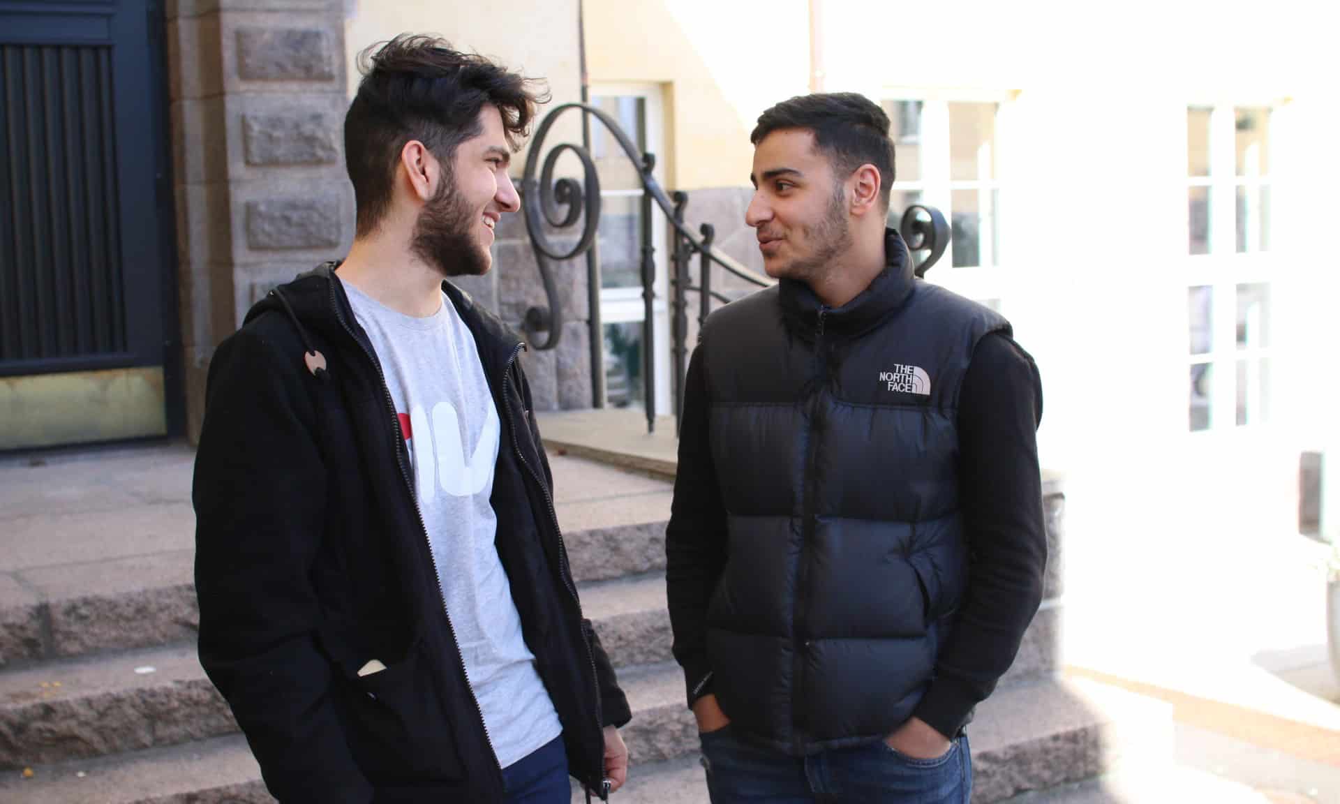 To unge gutter med mørkt hår og skjeggvekst står vendt mot hverandre i samtale. Smiler. En med mørk hettejakke, den andre i mørk genser med blå boblevest over. Foto SOS-barnebyer