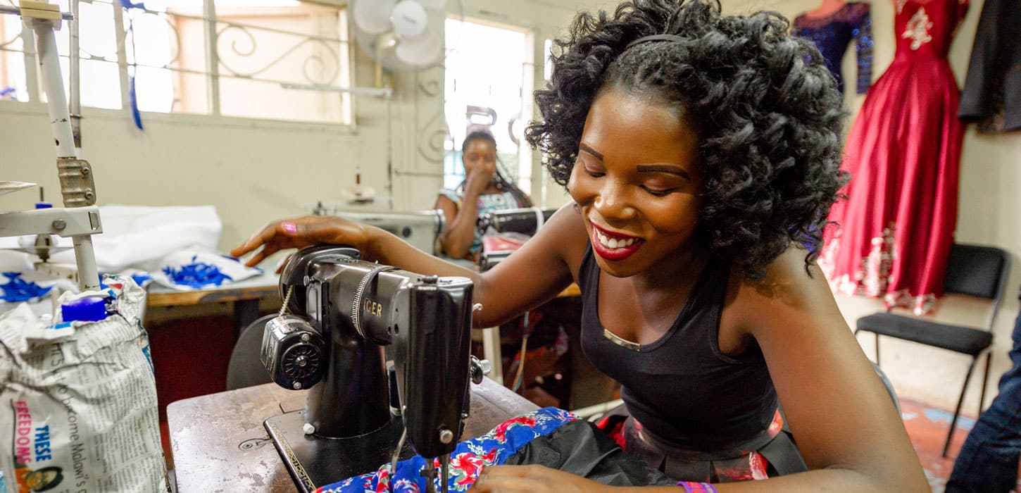 Malawi: Thoko sitter og syr en fargerik kjole i rødt hvit og blått, med en svart Singer symaskin. Hun har halvlangt krøllete hår, svart kortermet topp og smiler. Foto: Cornel van Heerden
