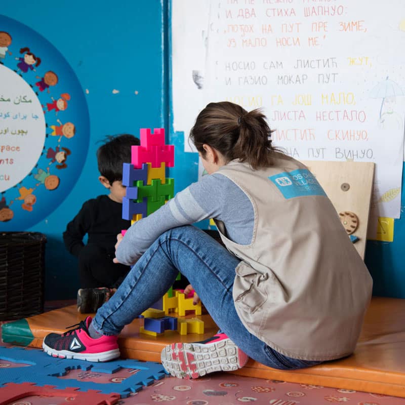 En medarbeider fra SOS-barnebyer med mørkt hår i hestehale, lyseblå genser, jeans og beite vest med SOS-barnebyers logo på ryggen sitter på gulvet med ryggen til og leker med en liten gutt med mørkt hår og mørk genser. Bildet er fra ett av SOS-barnebyers sosialsentre i Ukraina. Vårt arbeid i Europa. Foto: Katerina Ilievska