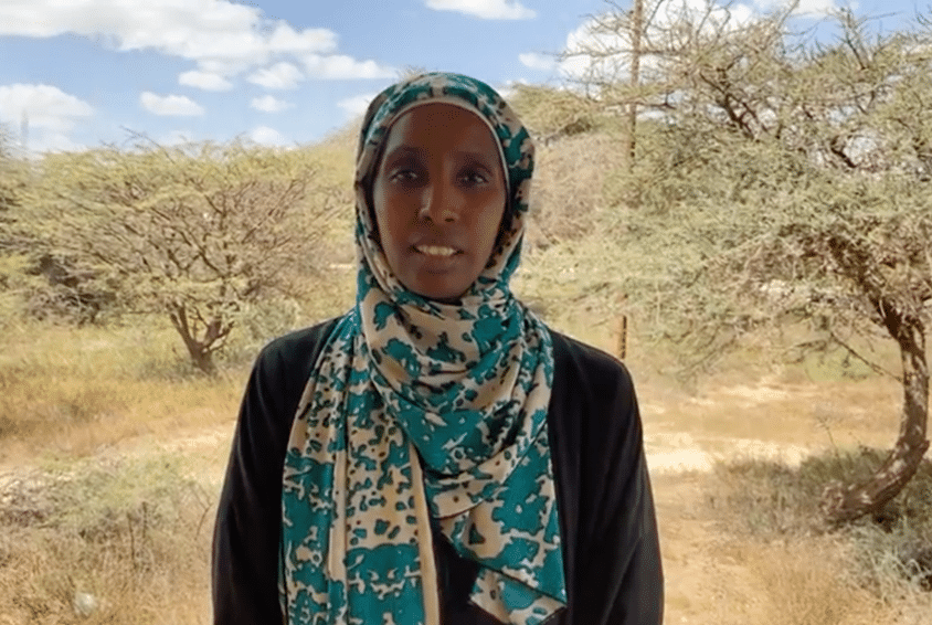 Sudan: Deqa Dimbil, SOS-barnebyers nødhjelpskoordinator i området. hun har beige og turkis hijab og mørk bluse. hun står ute blant trær og tørr gresskledd bakke. Foto: SOS-barnebyer