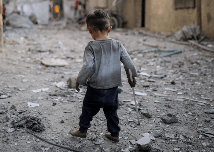 Gaza: Liten gutt med mørkt, kort hår, gra genser, svart joggebukse går barbeint i ei gate med knuste betongbiter. Bildet av ham er tatt bakfra. Foto: Belal Khaled