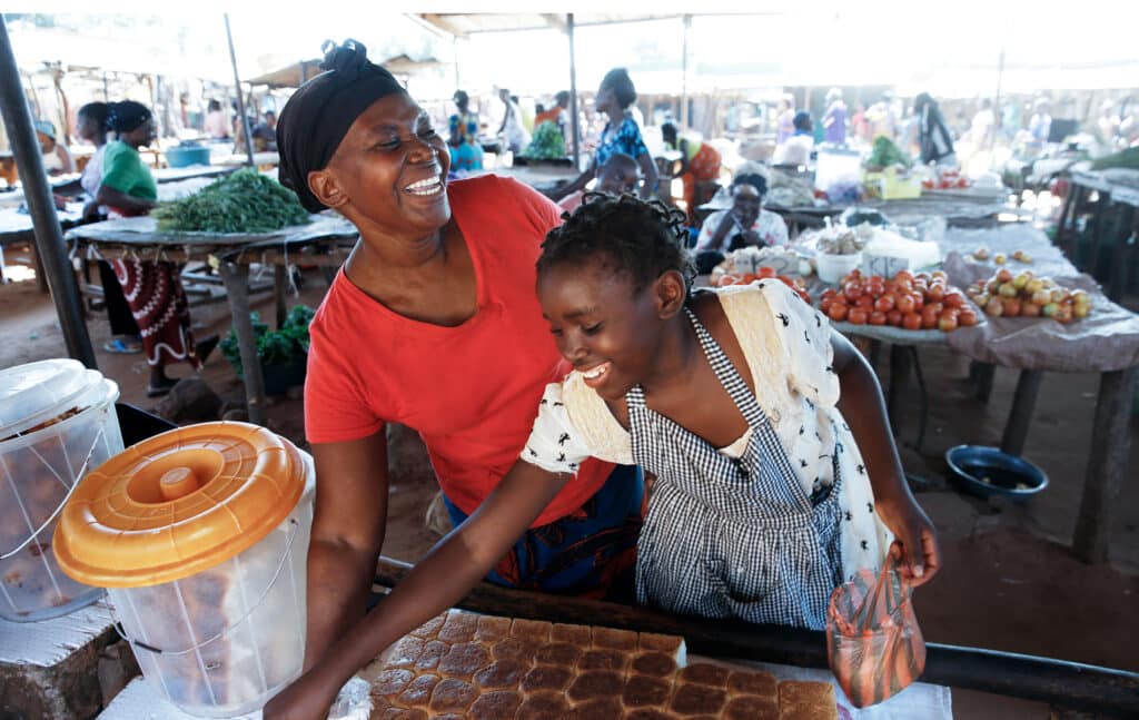 Babette har lille Noah i armene. Støtten Babette nå får gjennom SOS-barnebyers familieprogram i Zambia, gjør at hun nå klarer å studere, og være mor for lille Noah. Foto: Paul Audestad Moren til Betty i rød t-skjorte og skaut og blomstrete skjørt, står i salgsboden med et stort bredd med boller foran seg. Betty står ved siden av, hun har lys blomstrete kjole og lyseblått forkle. Begge smiler. Foto: Paal Audestad
