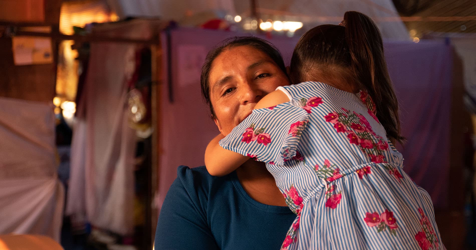 Bolivia: Hilda, med mørkt hår i hestehale og blå genser, holder den yngste datteren på armen. Hun har ryggen til, mørkt hår i hestehale og hvit og blå stripte kjole med rød blomster. Foto Monica Garcia