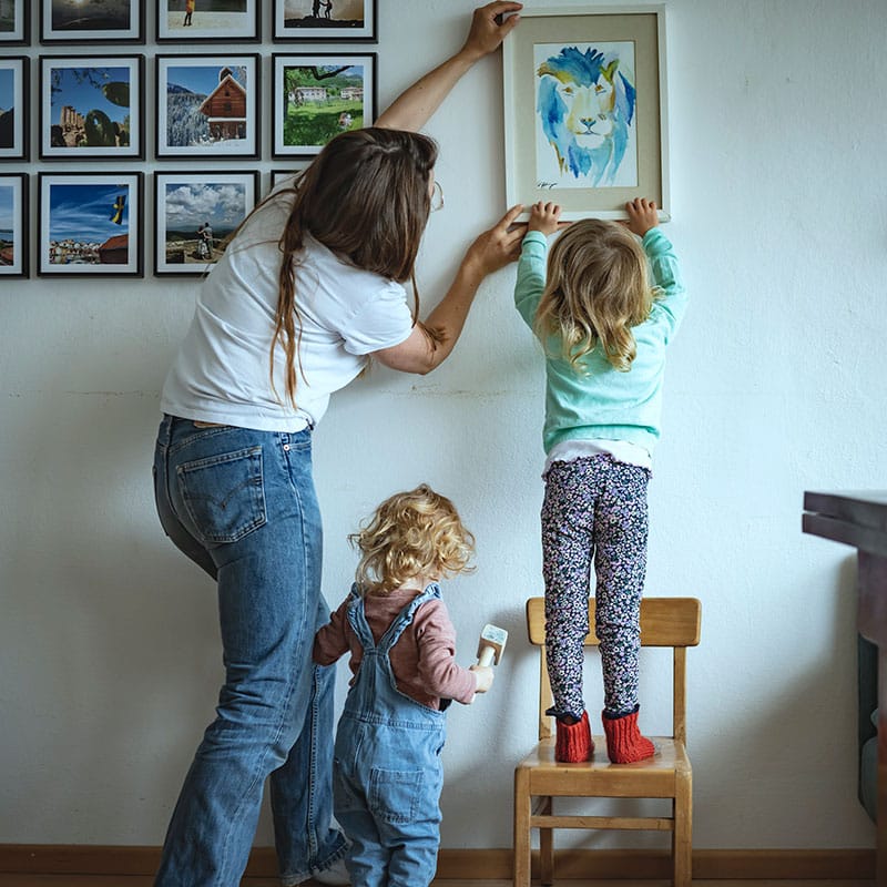 En mor med langt mørkeblondt hår, hvit t-skjorte og jeans hjelper datteren som står på en stol å henge opp et bilde. hun har blondt halvlangt hår, turkis genser og mønstrete bukse. Ved siden av står lillesøsteren med rose genser og selebukser. Foto Gerhard BErger